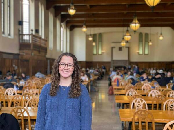 A student wearing glasses and a blue sweater smiles in the foreground of a busy dining hall. Wooden tables and chairs fill the space, and other students are seated in the background. Warm light from the chandeliers above illuminates the room.