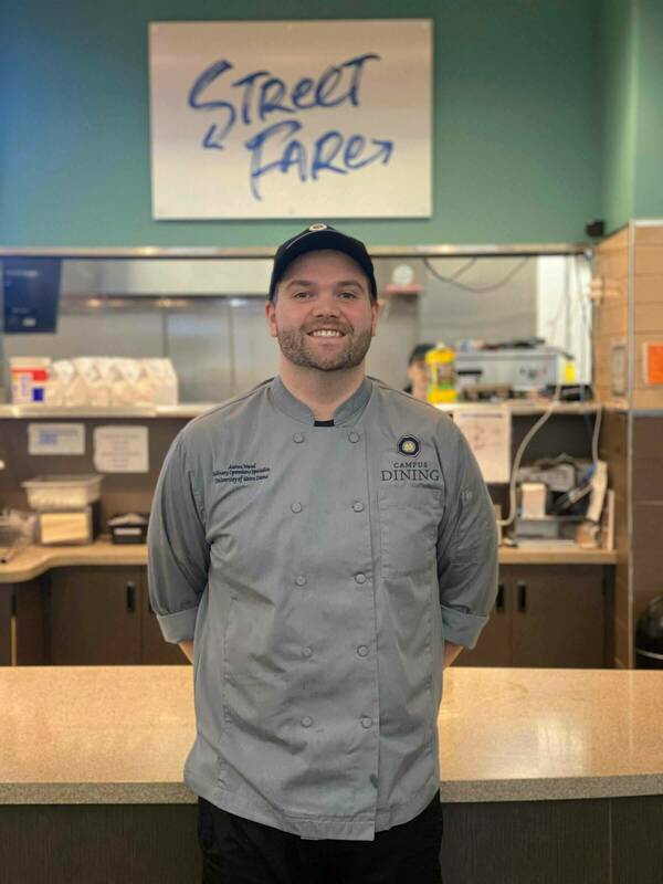 A chef in a gray chef's coat and black Notre Dame baseball cap stands with his hands behind his back and smiles at the camera. His name tag reads "Aaron Wood, Culinary Operations Specialist, University of Notre Dame." Behind him is a counter, kitchen equipment, and a sign reading "Street Fare".