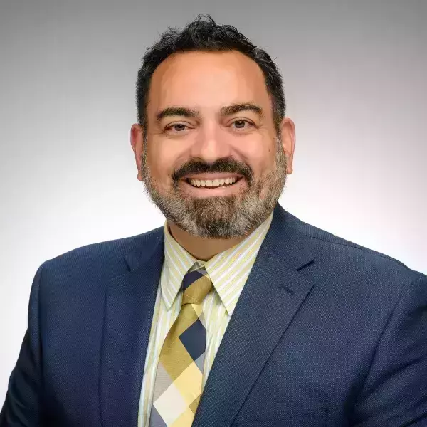 A man, wearing a navy blue suit and checkered tie, smiles for a headshot. A grey background encompasses the photo.