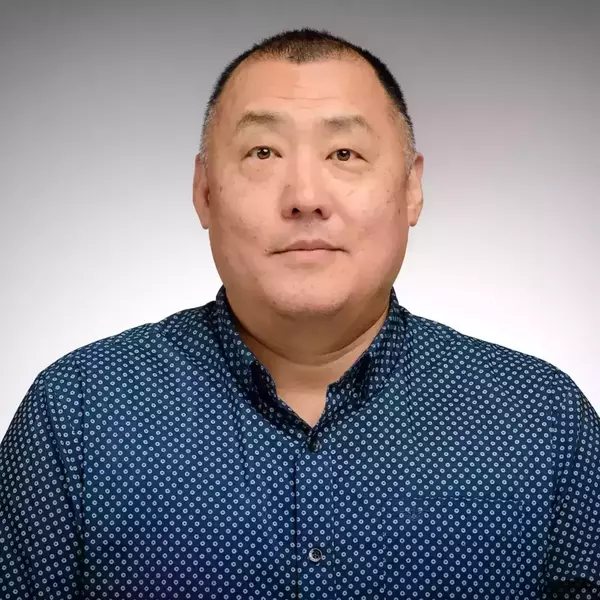 A man, wearing a navy and white polka-dot shirt, smiles for a headshot. A grey background encompasses the photo.