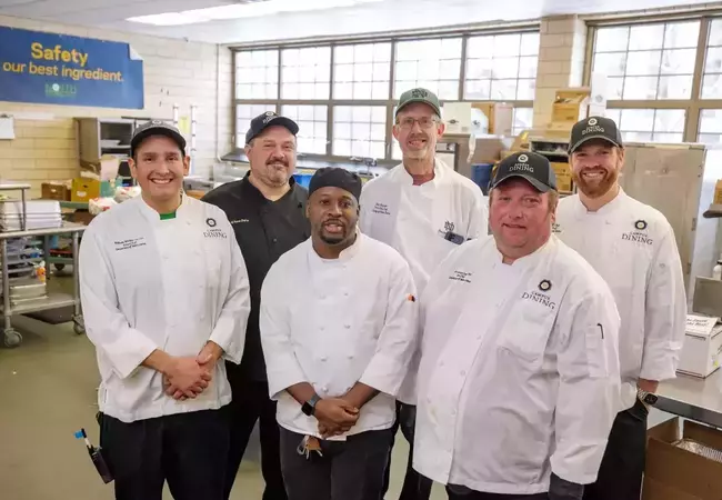 A group of six men, wearing chef coats and hats, smile for a picture in a commercial kitchen. Multiple pans and food, as well as a navy blue banner sign reading "Safety is our best ingredient" can be seen in the background.