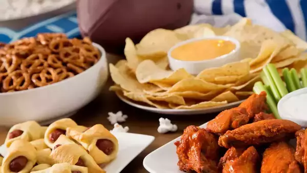 Super bowl party food featuring dishes like chips and dip, pigs in a blanket, pretzels, wings, and popcorn. A football sits behind the dishes.