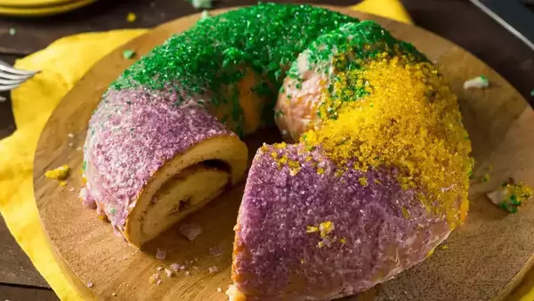 A circular king cake with purple, gold and green sprinkles sits on a wooden plate.