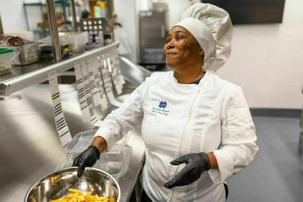 A woman, wearing a white chef coat and a white chef hat, smiles as she shakes a bowl of fries. Order tickets, additional supplies, and a commercial kitchen can be seen in the background.