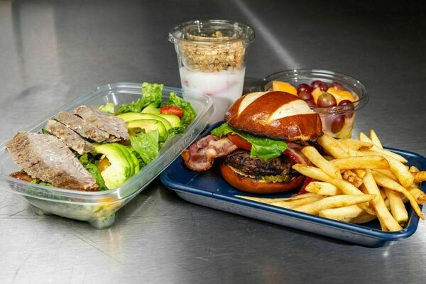 A colorful spread of multiple dishes of food including things like a burger, fries, fruit, salad, yogurt etc. All dishes sit on a silver commercial kitchen table.