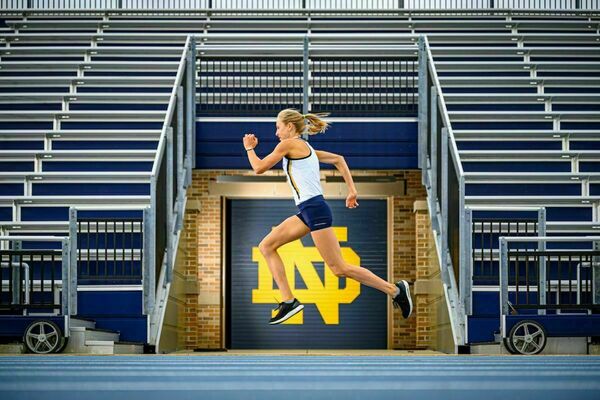 A women track runner is photographed in a running position in the air with the Notre Dame branded track and stadium stairs in the background.