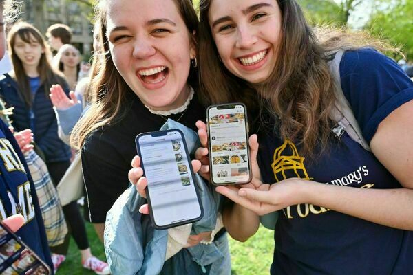 Two women stand and smile at the camera while holding up their phone screens, depicting the Grubhub mobile app ordering system. A sunny outside day and additional students fill the background.