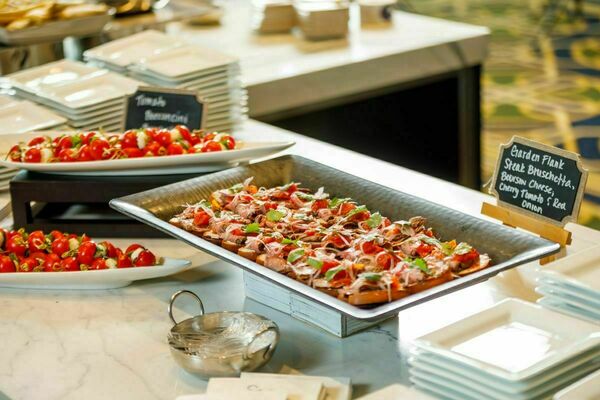 Multiple elaborate and neat catering dishes on a white marble table. Little bites like bruschetta and tomatoes fill the silver plates.