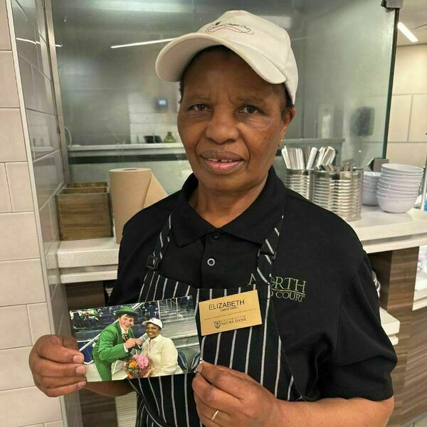 Elizabeth Ndani, a north dining hall attendant wearing a black shirt and a black and white pin stripe apron and a white hat, smiles for a picture in front of a serving station. She holds a printed photograph of herself and the Notre Dame Leprechaun.