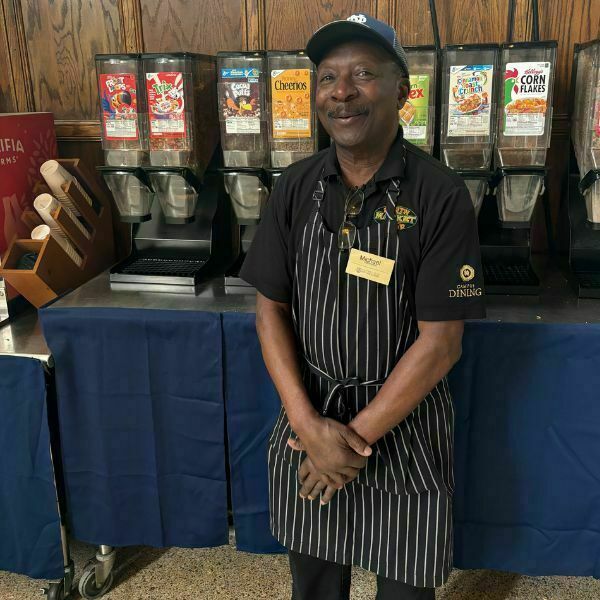 Michael, wearing a black and white striped apron, stands and smiles for a photo in front of the cereal station in South Dining Hall.