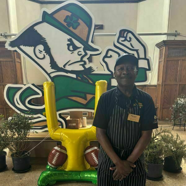 Michael, wearing a black and white striped apron, smiles for a photo in front of a life-size Notre Dame Leprechaun. Super Bowl decorations fill the area.