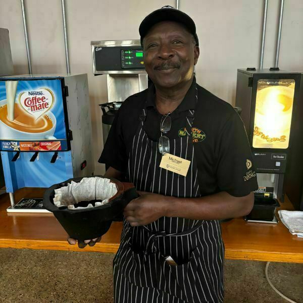 Michael, wearing a white and black striped apron, smiles as he holds coffee grinds in front of coffee machines in South Dining Hall.