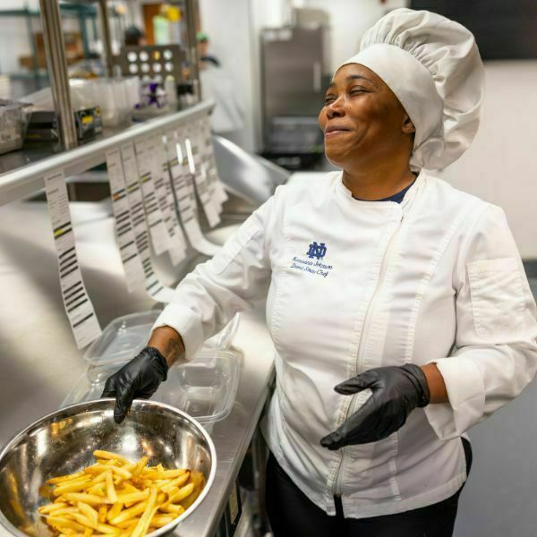 A chef, wearing a white chef coat and hat, smiles as she tosses an aluminum bowl of fries. Order receipts can be seen in a line as she prepares orders in a commercial kitchen.