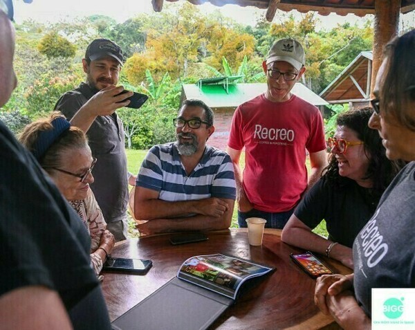 Multiple people stand and sit around an outdoor table with a coffee farm view in the background. One man is taking a top down photo of a photobook open on the table.