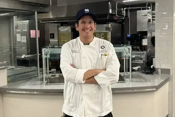 Chef William stands with his arms crossed and smiles for a photo. He wears a white chef coat and  blue hat. A commercial kitchen and serving station fills the background.