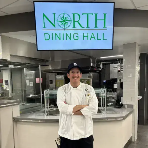Chef William stands with his arms crossed and smiles for a photo. He wears a white chef coat and blue hat. A commercial kitchen and serving station fills the background.
