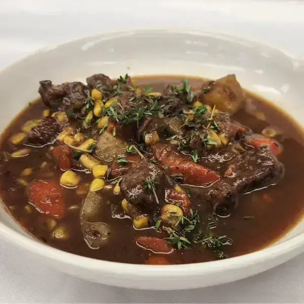 Close up view of bison stew in a white bowl amongst a white background. Items like bison meat, carrots, corn, rosemary and more can be seen.