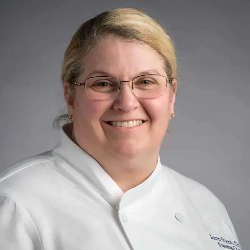 Headshot of Chef Laura Wiese, wearing a white chef coat, against a grey background.