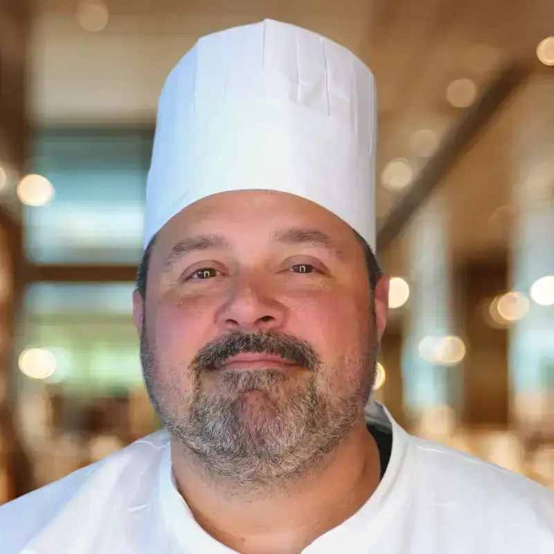 Chef Greg, wearing a white chef coat and hat, smiles for a headshot. Blurred lights fill the background.