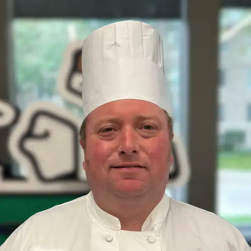 Chef Kris, wearing a white chef coat and hat, smiles for a headshot. A wooden statue of the Notre Dame Leprechaun and large windows fill the area behind him.