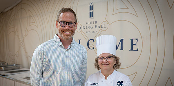 Assistant Professor of Economics Patrick Turner, a LEO researcher, and North Dining Hall’s Executive Chef Laura Wiese make up Notre Dame's YMCA chef team. They stand in front of a background of the South Dining Hall logo and Chef Laura wears a white chef coat and hat.