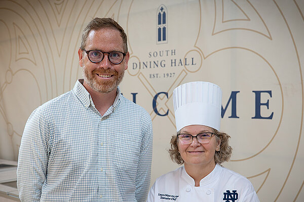 Assistant Professor of Economics Patrick Turner, a LEO researcher, and North Dining Hall’s Executive Chef Laura Wiese make up Notre Dame's YMCA chef team. They stand in front of a background of the South Dining Hall logo and Chef Laura wears a white chef coat and hat.
